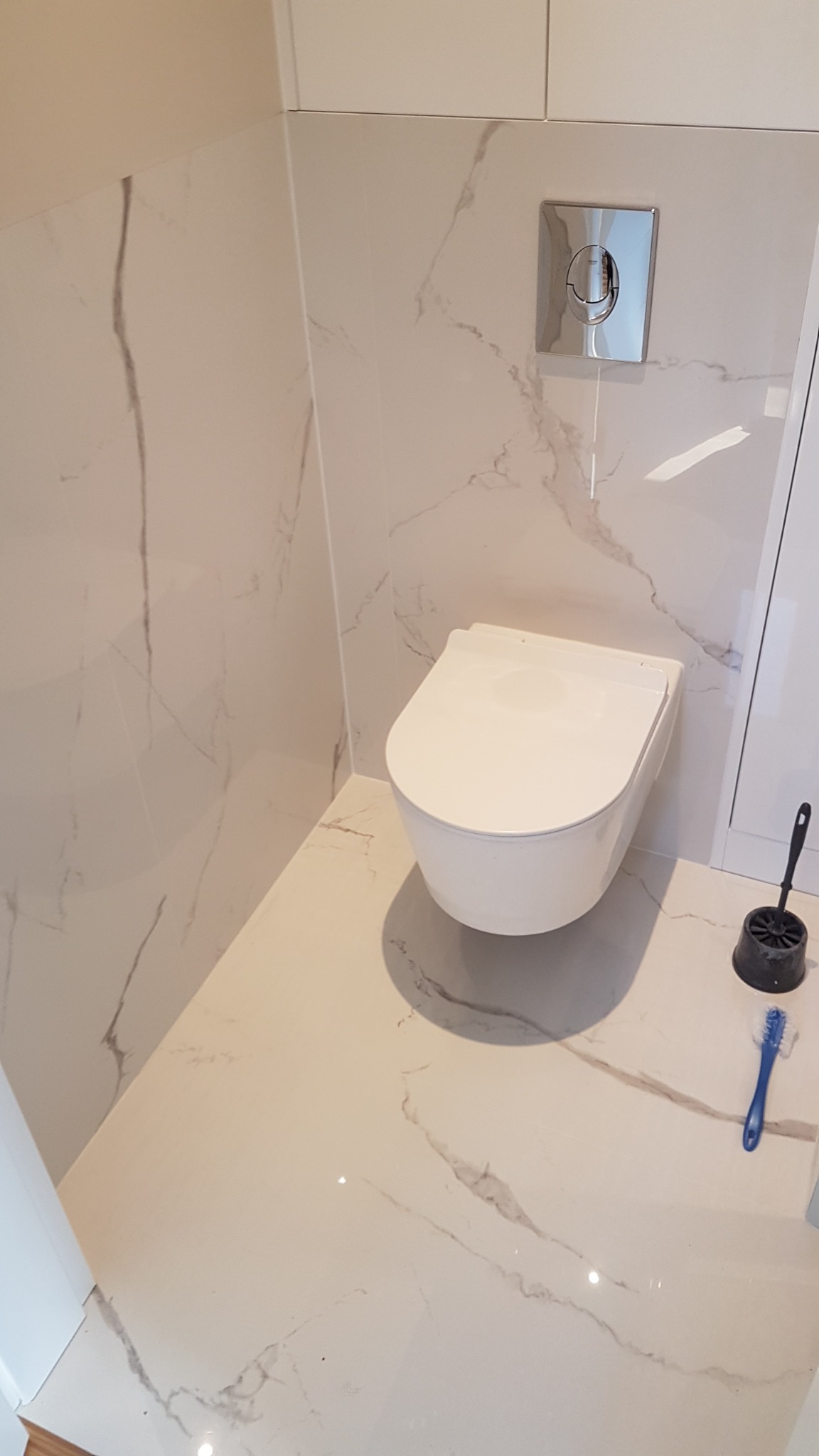 Nowoczesna, podwieszana toaleta w łazience wykończonej płytkami imitującymi marmur, z chromowanym przyciskiem spłukującym i akcesoriami toaletowymi.