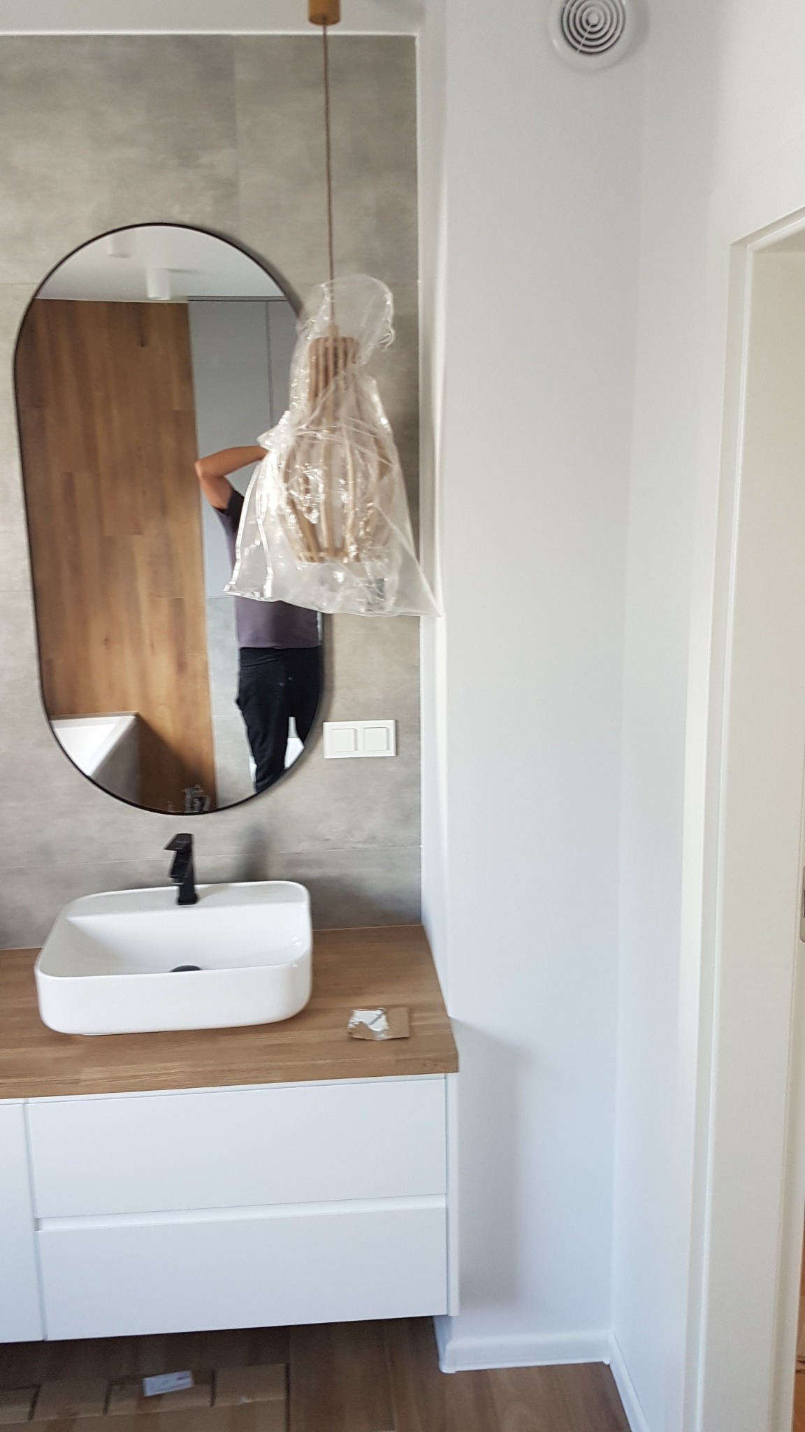 Nowoczesna łazienka z umywalką nablatową, owalnym lustrem i lampą wiszącą w folii ochronnej, szare płytki na ścianie.