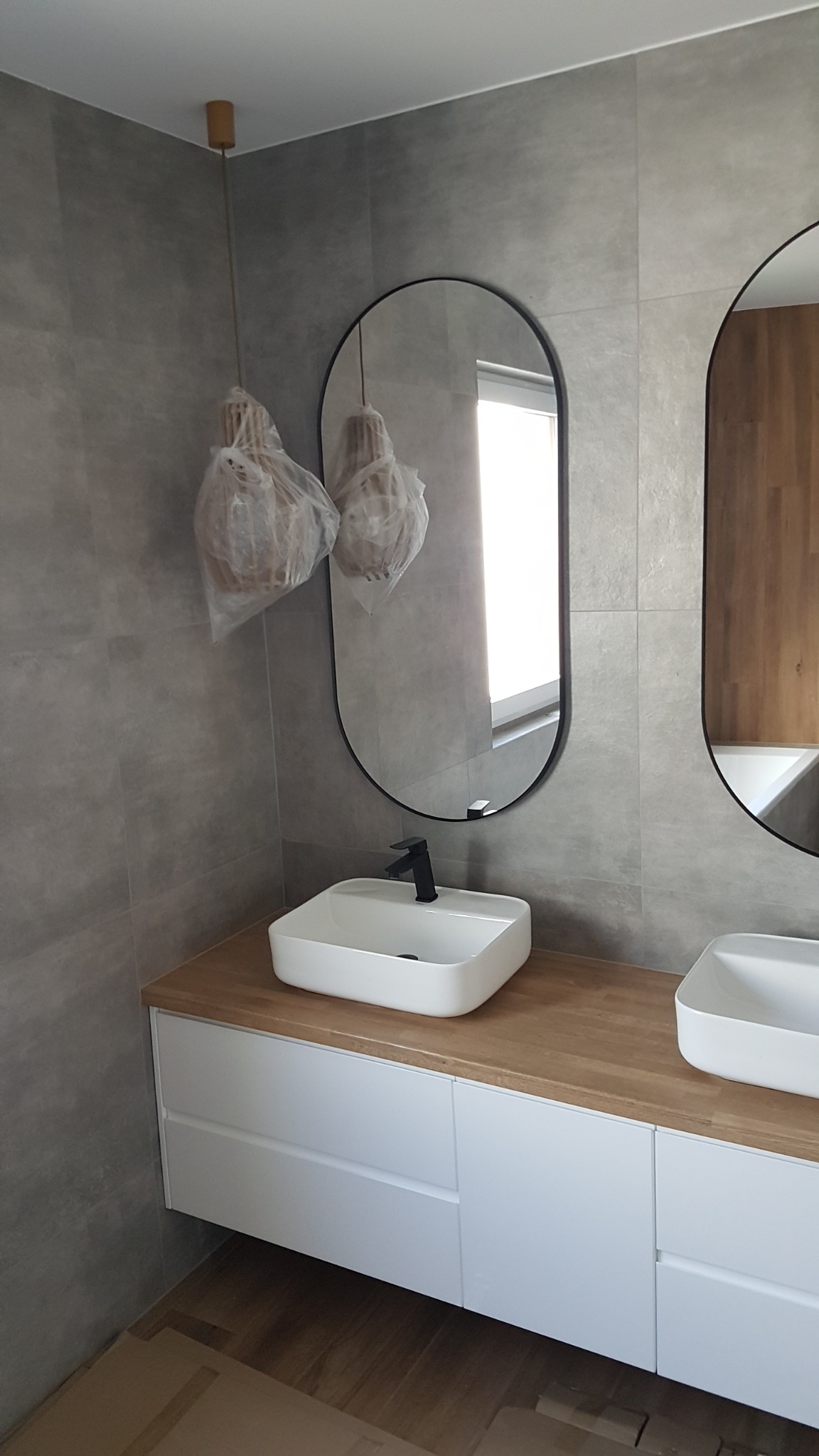 Nowoczesna łazienka w szarościach z dwiema owalnymi umywalkami nablatowymi i lustrami w czarnych ramach, wiszące lampy w osłonach ochronnych, drewniany blat i białe szafki pod umywalkami.