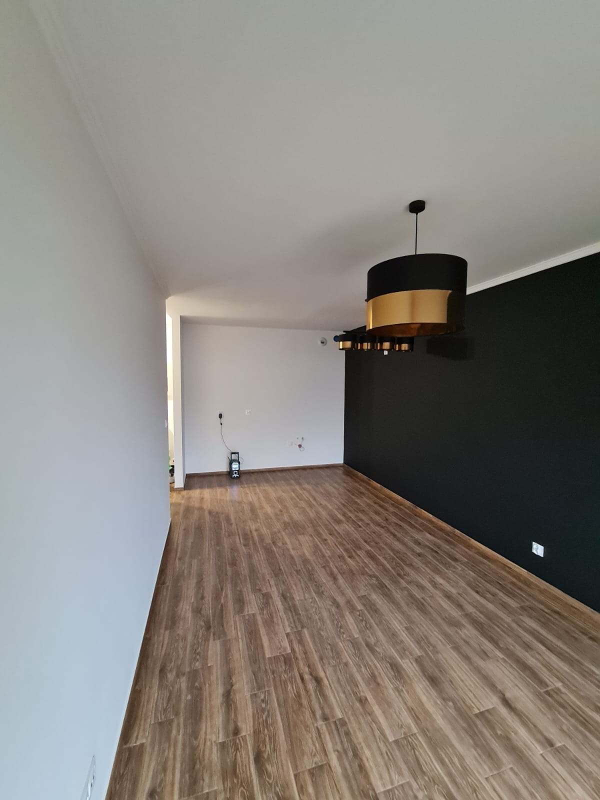 Pomalowane na biało i czarno ściany w pustym pokoju z panelami podłogowymi, widoczny nowoczesny żyrandol w kolorze czarno-złotym i poziomica laserowa.