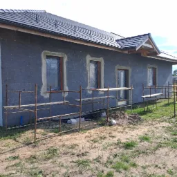 Dom jednorodzinny w trakcie ocieplania elewacji, widoczne rusztowania, okna zabezpieczone folią, ściany pokryte szarym materiałem izolacyjnym, przygotowane pod dalsze prace wykończeniowe.