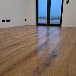 Nowo położona, drewniana podłoga w jasnym pomieszczeniu z dużymi, przeszklonymi drzwiami balkonowymi, przez które widać zimowy krajobraz.