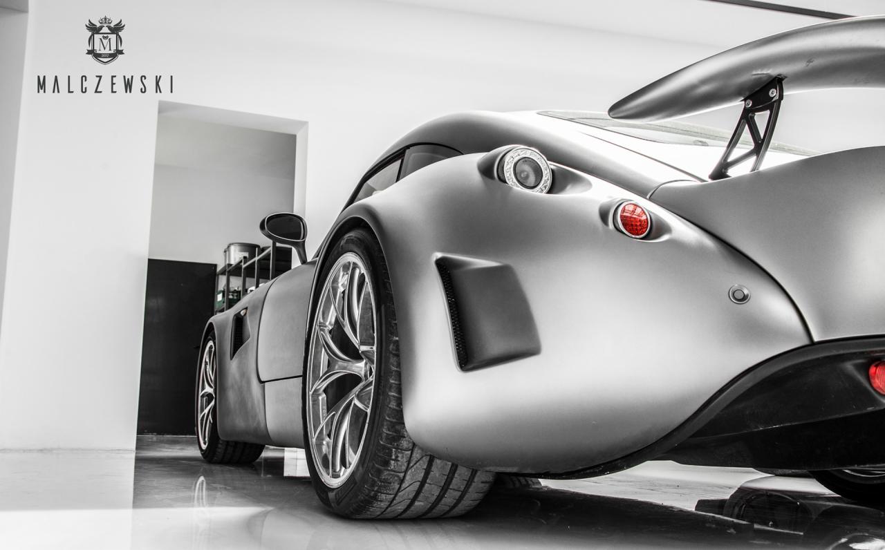 Tył matowego, srebrnego sportowego auta z dużym spojlerem i logo firmy Malczewski na ścianie, w jasnym studiu.