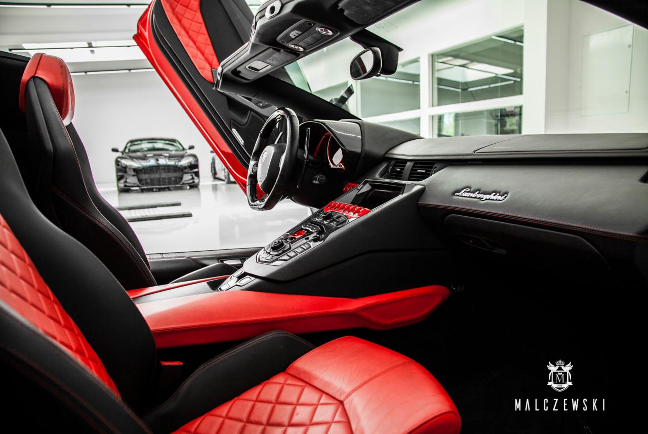 Wnętrze luksusowego samochodu sportowego Lamborghini z czarno-czerwonymi skórzanymi siedzeniami, widok z boku z otwartymi drzwiami, na zewnątrz zaparkowany Aston Martin