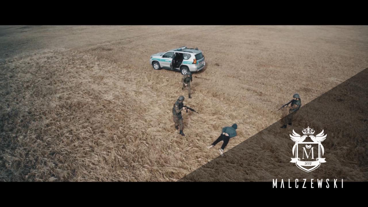 Scena z filmu akcji: żołnierze z bronią obezwładniają mężczyznę na polu, w tle radiowóz policyjny z otwartymi drzwiami.