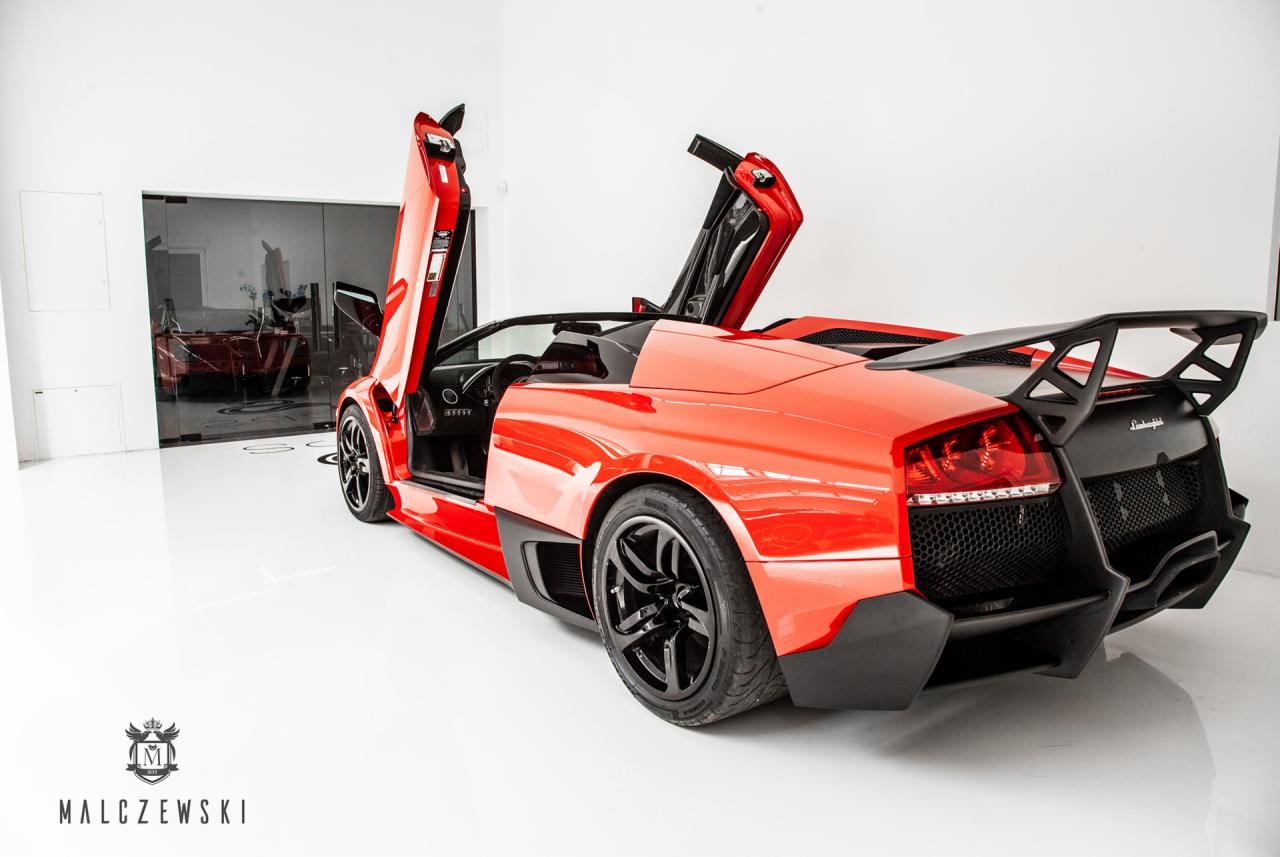 Błyszczący, czerwony, sportowy kabriolet z otwartymi drzwiami w stylu nożycowym, zaparkowany w minimalistycznym, białym garażu z widocznym logo firmy Malczewski.