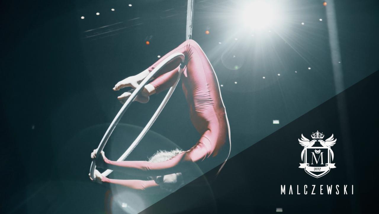 Akrobatka w czerwonym kostiumie wykonuje skomplikowaną figurę na białym kole cyrkowym, oświetlona reflektorami, w tle logo firmy.