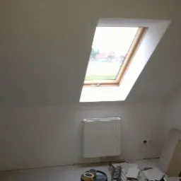 Poddasze po remoncie: białe ściany, okno dachowe z drewnianą ramą, grzejnik, widoczne materiały budowlane i narzędzia na podłodze.