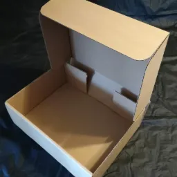 Otwarta, pusta, brązowa, tekturowa skrzynka z wewnętrznymi przegródkami, widoczna na ciemnym, pogniecionym tle.