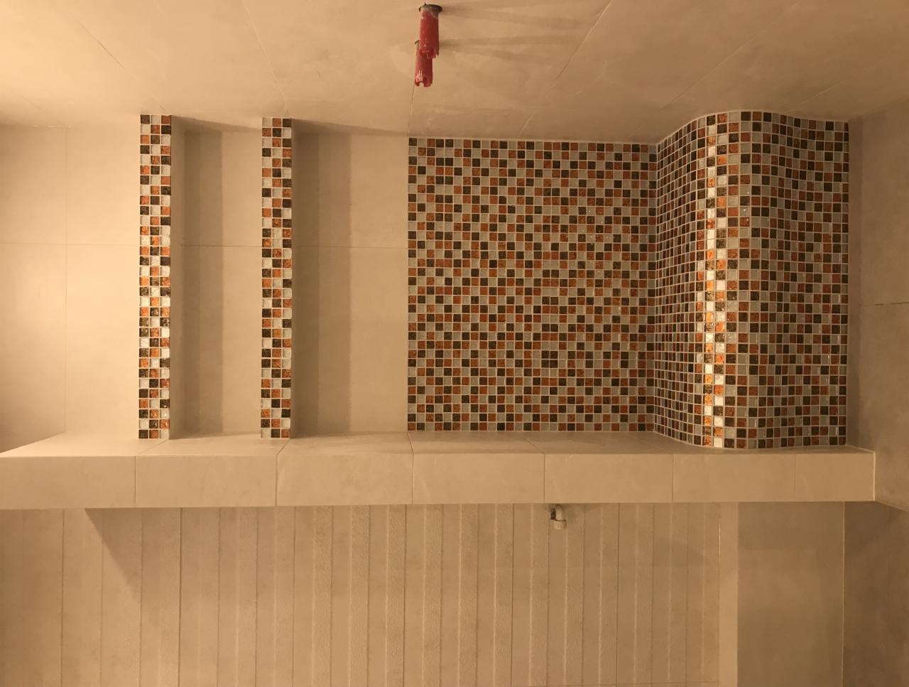 Wykończona ściana w łazience z mozaiką w odcieniach brązu, beżu i bieli, ułożoną w pasy i na zaokrąglonym narożniku, z widoczną półką i pionowymi panelami z płytek.
