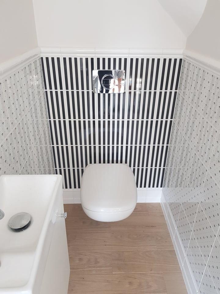 Nowoczesna toaleta z białymi płytkami w drobne wzory i czarno-białymi paskami na ścianie za muszlą, drewniana podłoga, biała umywalka.
