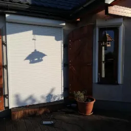 Biała roleta zewnętrzna częściowo opuszczona na tle drewnianych okiennic i okna, z widocznym cieniem karmnika dla ptaków.