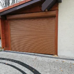 Brązowa, pozioma osłona okienna garażu w drewnianej obudowie, częściowo opuszczona, widoczna kostka brukowa i fragment drugiego garażu z samochodem w środku.