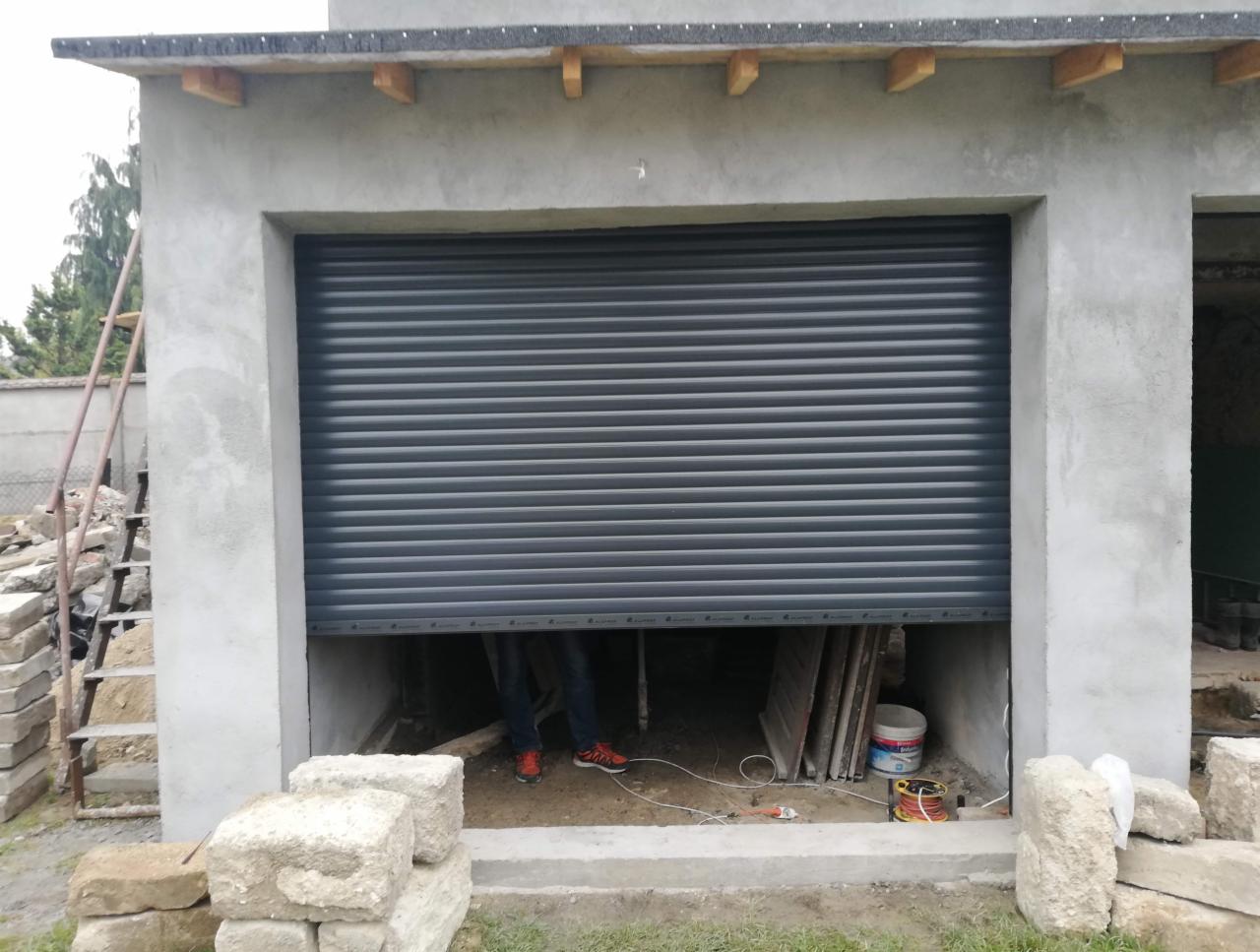 Montaż szarej rolety zwijanej w garażu, widoczne przewody elektryczne, cegły i drabina, osoba w jeansach i czerwonych butach wewnątrz garażu.