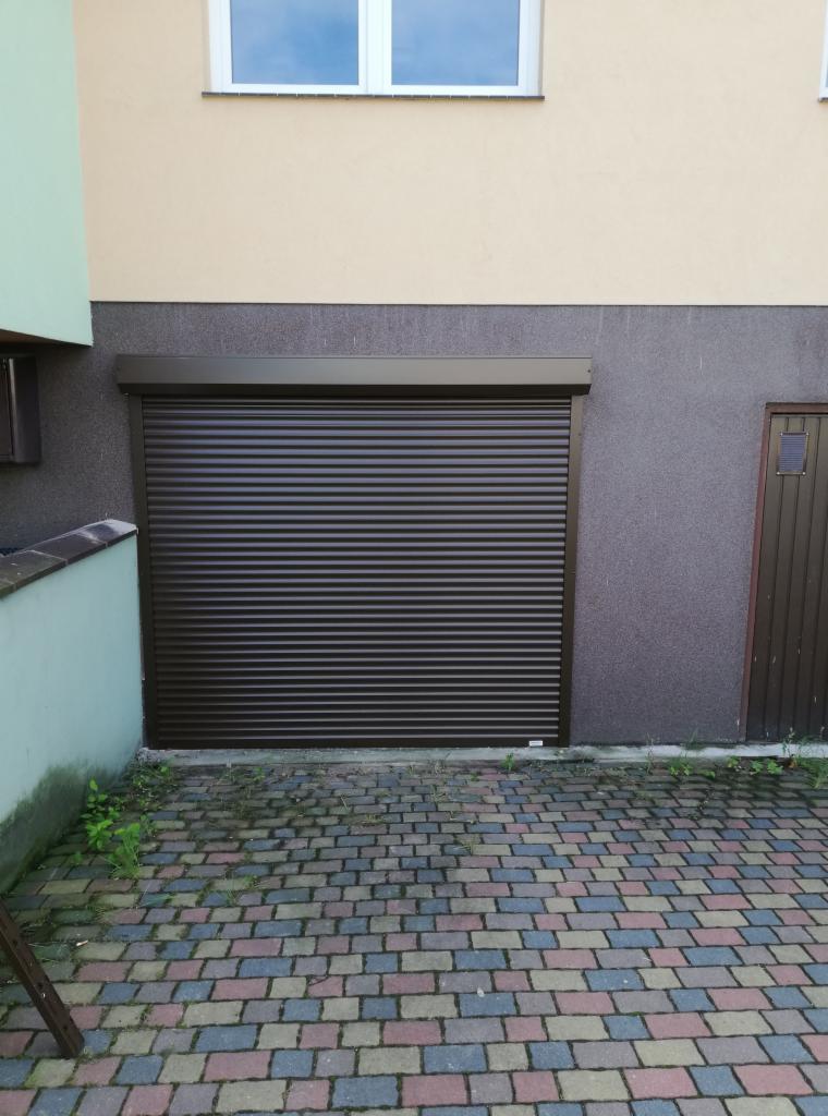 Zewnętrzna, brązowa roleta zwinięta na całą wysokość okna garażowego, widoczna na tle budynku z ceglaną kostką brukową.