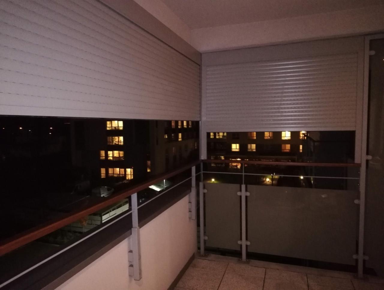 Zewnętrzne osłony okienne częściowo opuszczone na balkonie, widok nocny na oświetlony budynek naprzeciwko.