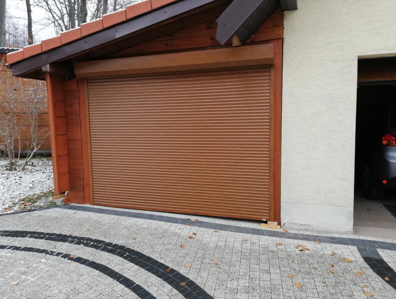 Brązowa, pozioma osłona okienna garażu w drewnianej obudowie, częściowo opuszczona, widoczna kostka brukowa i fragment drugiego garażu z samochodem w środku.