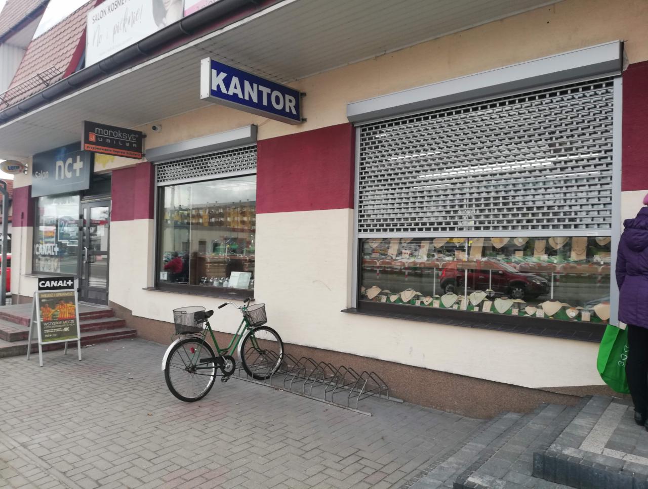 Fasada budynku z dwoma oknami zabezpieczonymi metalowymi roletami, rower zaparkowany przed budynkiem, szyld 'Kantor' widoczny nad oknem po lewej stronie.