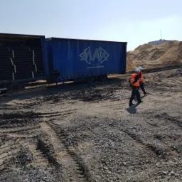 Dwaj pracownicy w kamizelkach odblaskowych przechodzą obok ciężarówki z otwartą naczepą wypełnioną czarnymi rurami, w tle hałda piasku i budynek.