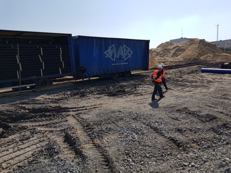 Dwaj pracownicy w kamizelkach odblaskowych przechodzą obok ciężarówki z otwartą naczepą wypełnioną czarnymi rurami, w tle hałda piasku i budynek.