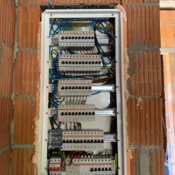 Rozdzielnica elektryczna zamontowana w ścianie z czerwonej cegły, widoczne liczne przewody i zabezpieczenia, uszczelniona pianką montażową.