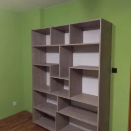 Pusta, drewniana regał na książki z licznymi przegródkami, wykonany z płyty w odcieniach szarości, stoi na drewnianej podłodze przy zielonej ścianie z widocznym gniazdkiem elektrycznym.