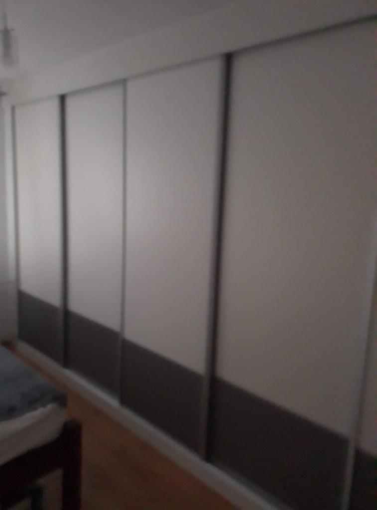 Czterodrzwiowa szafa przesuwna z białymi frontami i szarym dolnym panelem, widoczna z boku w słabym oświetleniu.