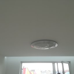 TOM-BUD - Biały sufit z okrągłą lampą sufitową, widok na miasto przez okno z białymi ramami.