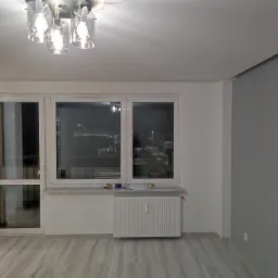 Wnętrze pokoju po remoncie: panele podłogowe w odcieniach szarości, białe ściany z grzejnikiem pod oknem, nowoczesna lampa sufitowa z przezroczystymi kloszami.