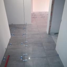 TOM-BUD - Układanie szarych płytek podłogowych z użyciem poziomicy i plastikowych krzyżyków dystansowych w korytarzu podczas remontu.