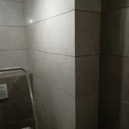 Ściana wyłożona podłużnymi, błyszczącymi płytkami w odcieniach szarości i beżu, z widoczną drabiną i taśmą malarską na jednym ze stopni.