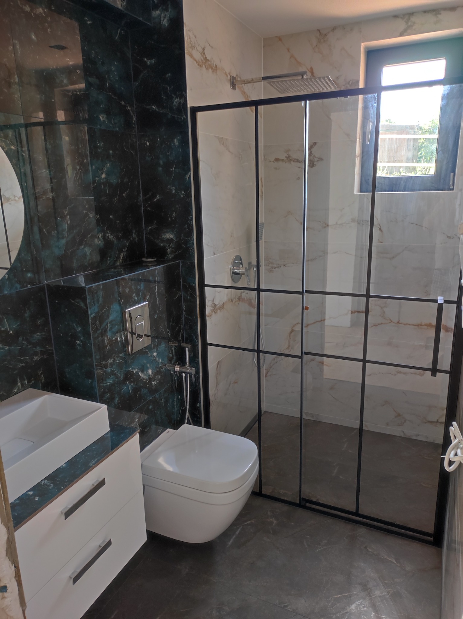 Nowoczesna łazienka z kabiną prysznicową w czarnej ramie, umywalką nablatową na białej szafce z szufladami, toaletą podwieszaną i płytkami imitującymi marmur w odcieniach bieli i zieleni.