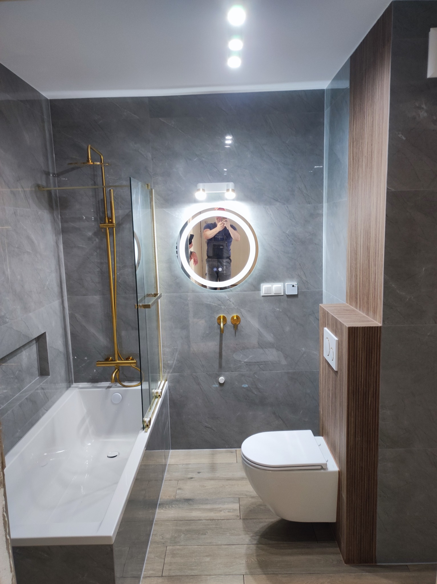 Wykończona łazienka: szare płytki imitujące marmur, złota armatura, okrągłe lustro z podświetleniem, podwieszana toaleta. Nowoczesny design z dbałością o detale.