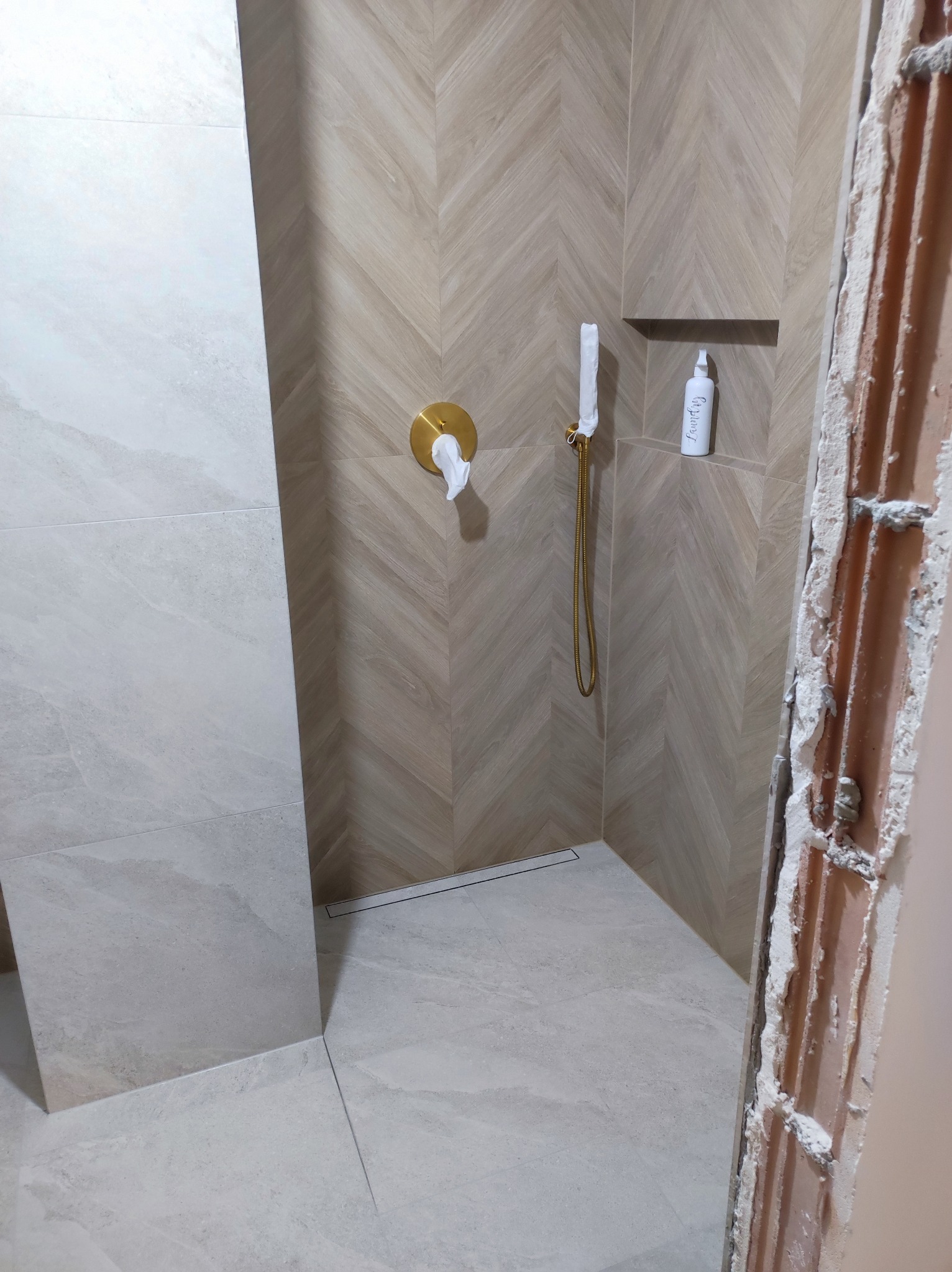 Wykończona kabina prysznicowa z beżowymi płytkami ułożonymi w jodełkę, złotą armaturą i wnęką na kosmetyki. Fragment surowej ściany z cegły.