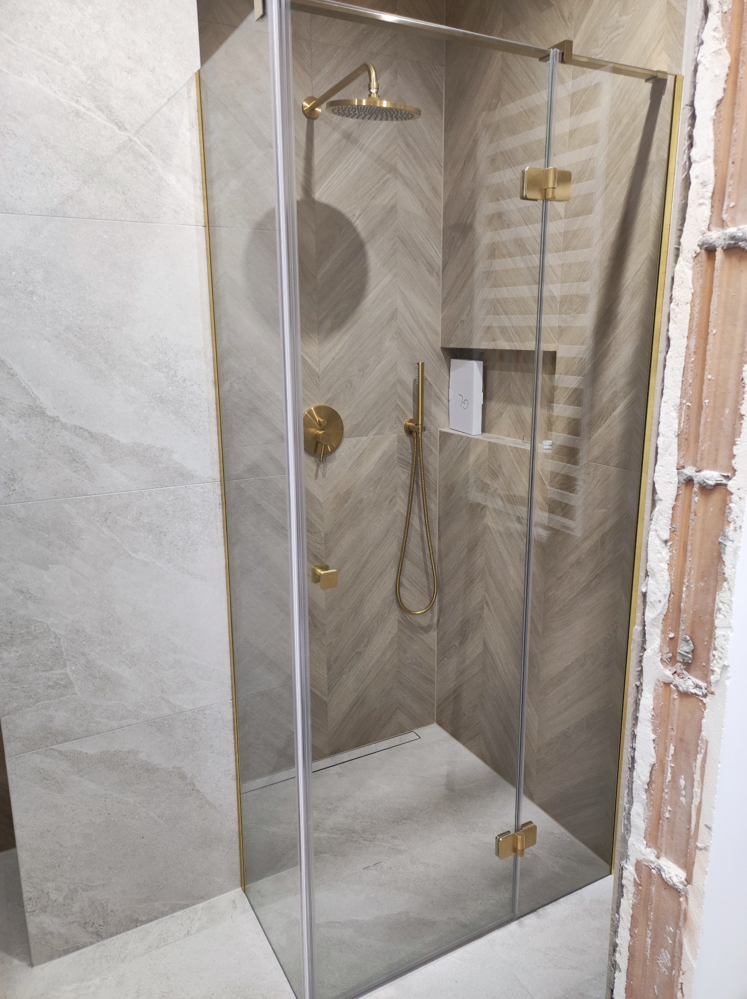 Kabina prysznicowa ze złotymi elementami na tle płytek w jodełkę. Widoczny fragment ściany w stanie surowym. Nowoczesny design łazienki.