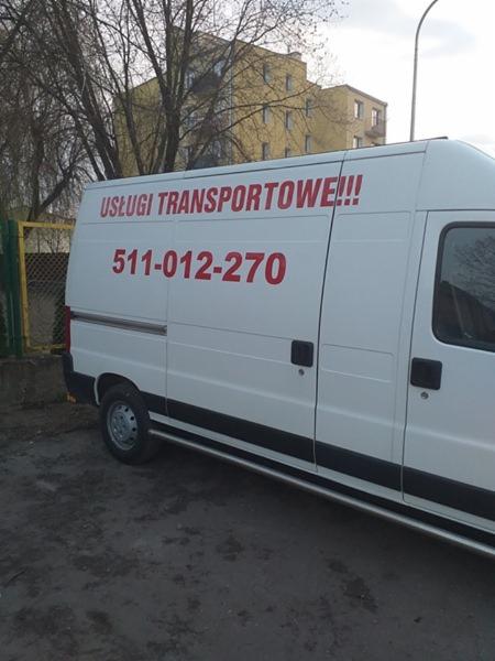 Biały bus z czerwonym napisem 'USŁUGI TRANSPORTOWE!!!' i numerem telefonu, zaparkowany na zewnątrz budynku.