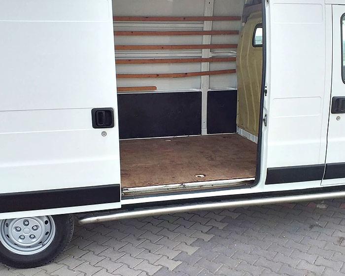 Wnętrze częściowo opróżnionego białego busa z drewnianymi półkami i brązową podłogą, widok z boku przez otwarte drzwi przesuwne.