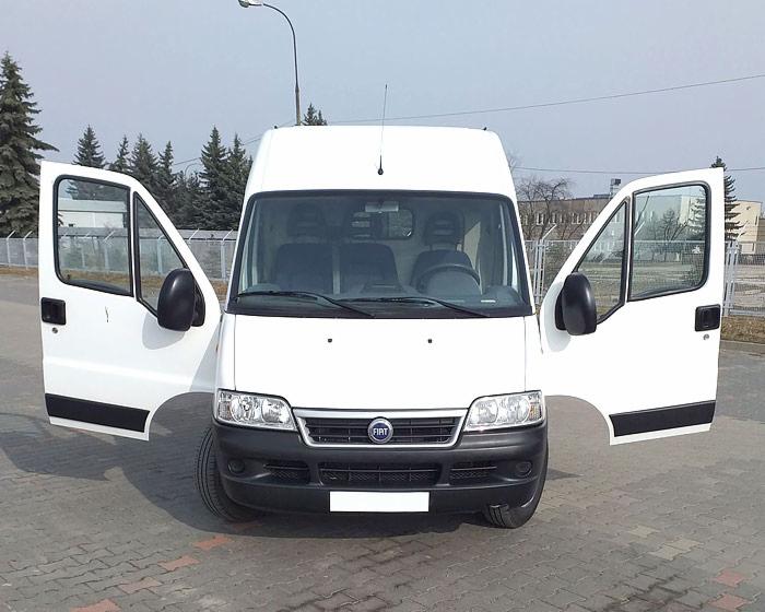 Biały bus Fiat Ducato z otwartymi drzwiami kabiny, zaparkowany na brukowanym placu, widok z przodu.