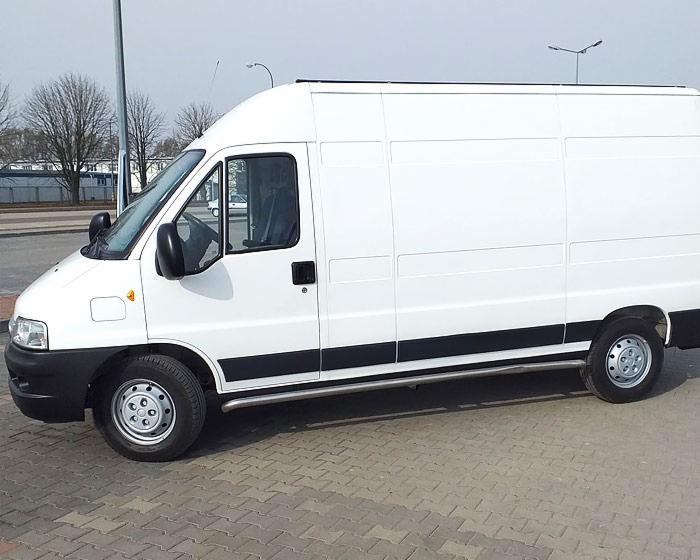 Biały, dostawczy bus z relingami bocznymi na tle szarej kostki brukowej i drzew bez liści.