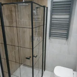 Nowoczesna kabina prysznicowa z czarnymi profilami i deszczownicą, grzejnik drabinkowy w szarym kolorze, płytki imitujące drewno na ścianie prysznica, biała ceramika łazienkowa.