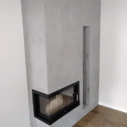 Nowoczesny kominek narożny z szarą, surową gładzią, czarnym obramowaniem wkładu i wnęką, widoczne drewnopodobne panele podłogowe.