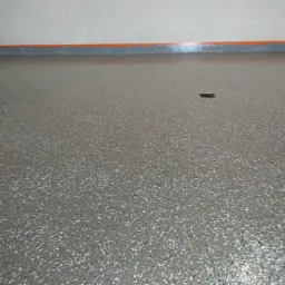 Szara, żywiczna posadzka z drobnymi, kolorowymi wtrąceniami, zabezpieczona taśmą malarską przy ścianie, z widocznym niewielkim elementem na powierzchni.