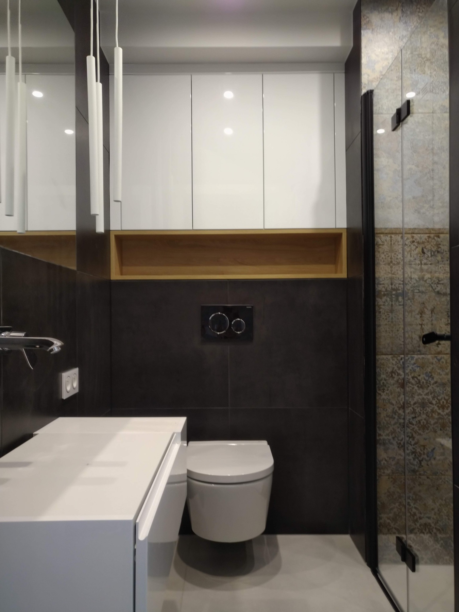 Nowoczesna łazienka z ciemnymi płytkami, białymi szafkami nad zabudowaną toaletą i kabiną prysznicową z dekoracyjnymi płytkami. Umywalka nablatowa z chromowaną baterią i minimalistycznym oświetleniem.