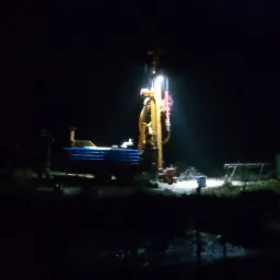 Żółta wiertnica oświetlona reflektorem podczas nocnych prac wiertniczych, widoczny fragment niebieskich rur i sylwetka pracownika w tle.