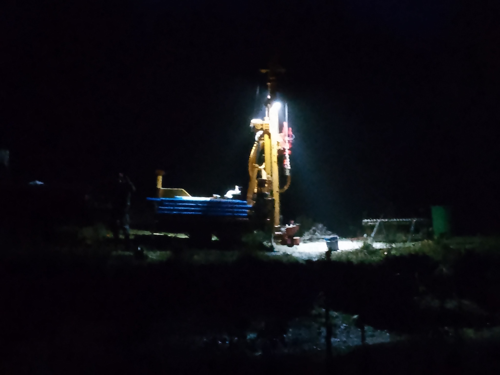 Żółta wiertnica oświetlona reflektorem podczas nocnych prac wiertniczych, widoczny fragment niebieskich rur i sylwetka pracownika w tle.
