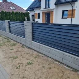 Nowoczesne ogrodzenie z poziomych, grafitowych paneli zamontowane na betonowych słupkach i podmur&oacute;wce, na tle nowego domu z jasną elewacją i brązowymi akcentami.