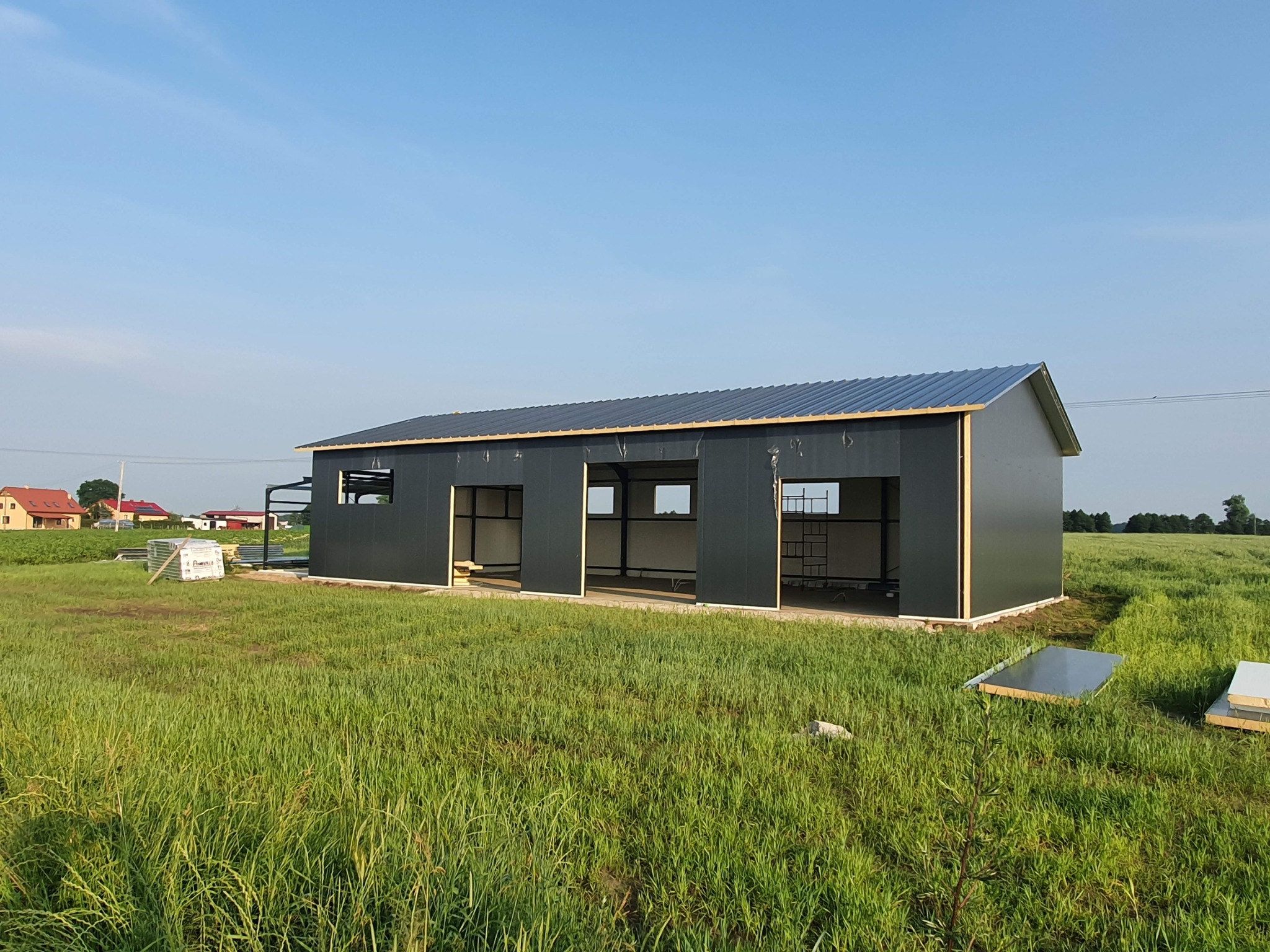 Budowa hali z metalową konstrukcją na zielonej łące w słoneczny dzień. Widoczne panele ścienne i dach z blachy. Częściowo zmontowana konstrukcja.