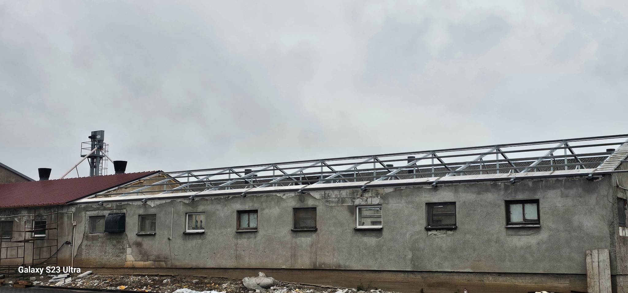 Szara hala w trakcie budowy dachu z metalową konstrukcją. Widoczne okna i fragment dachu z czerwoną dachówką. Pochmurne niebo w tle.