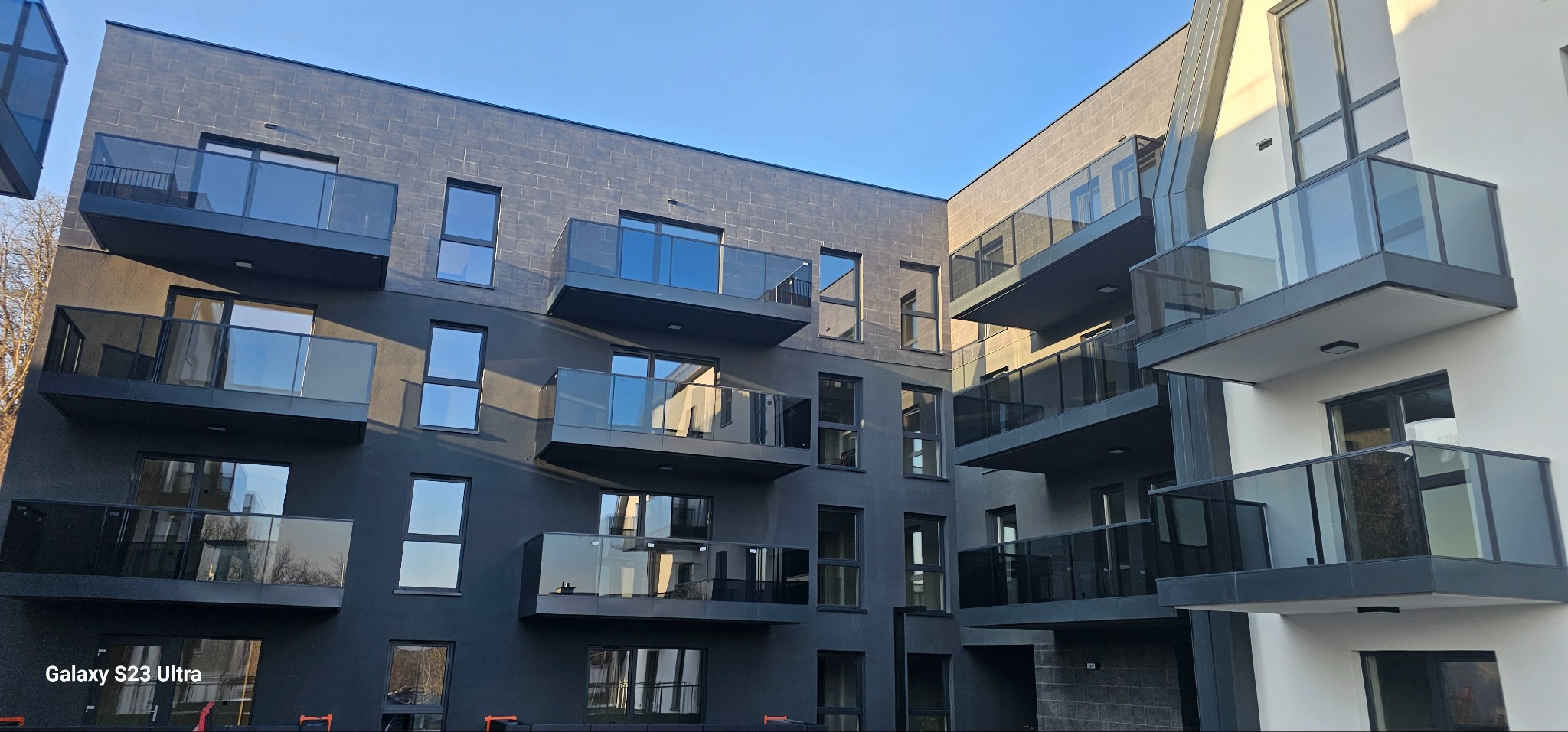 Nowoczesny budynek wielorodzinny z balkonami o szklanych balustradach i grafitowej elewacji. Widok na dziedziniec wewnętrzny w słoneczny dzień.