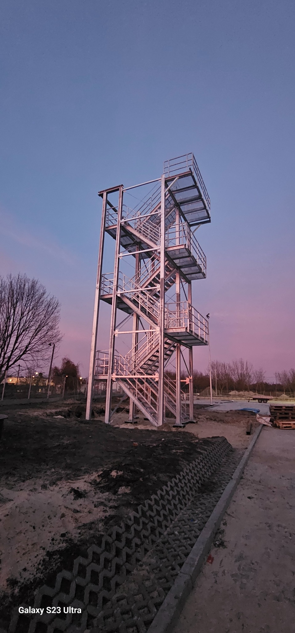 Wysoka, metalowa konstrukcja schodowa z platformami, wzniesiona na placu budowy, sfotografowana pod wieczornym, różowo-fioletowym niebem. Widoczne elementy krajobrazu.